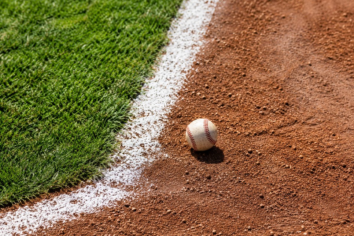 Overhead view of a baseball infield's white chalk lines and raked dirt under harsh midday sun, a lone baseball near the base path with heat shimmer rising.
