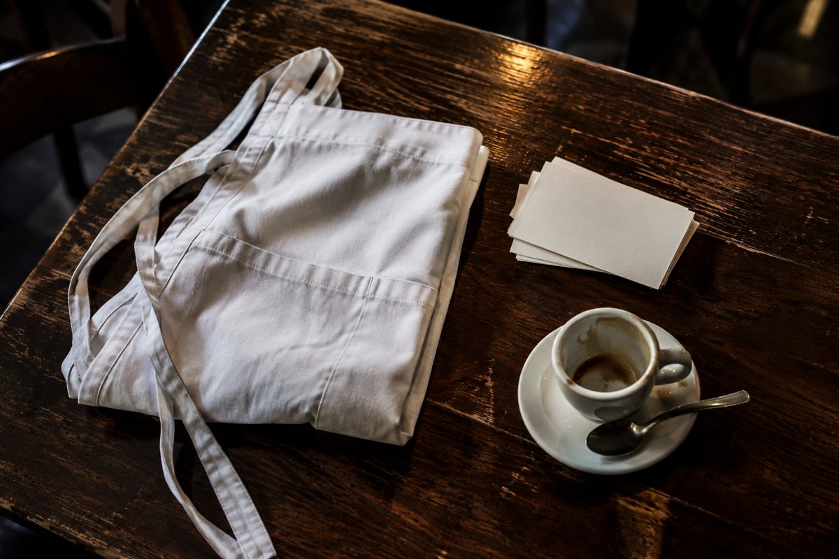 Overhead view of a worn wooden restaurant table with a folded white apron, a stack of blank reservation cards and an empty espresso cup, lit from behind in a dark dining room.