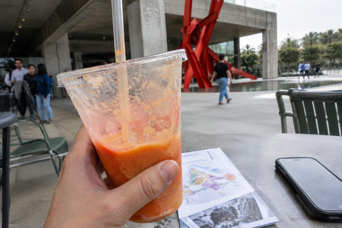 A hand holds an orange-pink smoothie at an outdoor cafe table beneath a concrete museum building on an overcast day, a painted steel sculpture blurred in the distance.