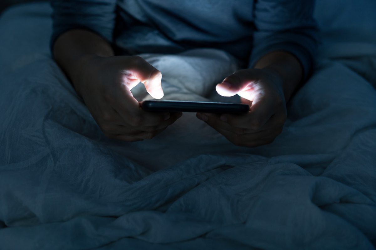 A teenager's hands holding a smartphone in a dimly lit room, face out of frame, screen glow lighting the fingers — illustrating FBI warnings about account takeover and sextortion