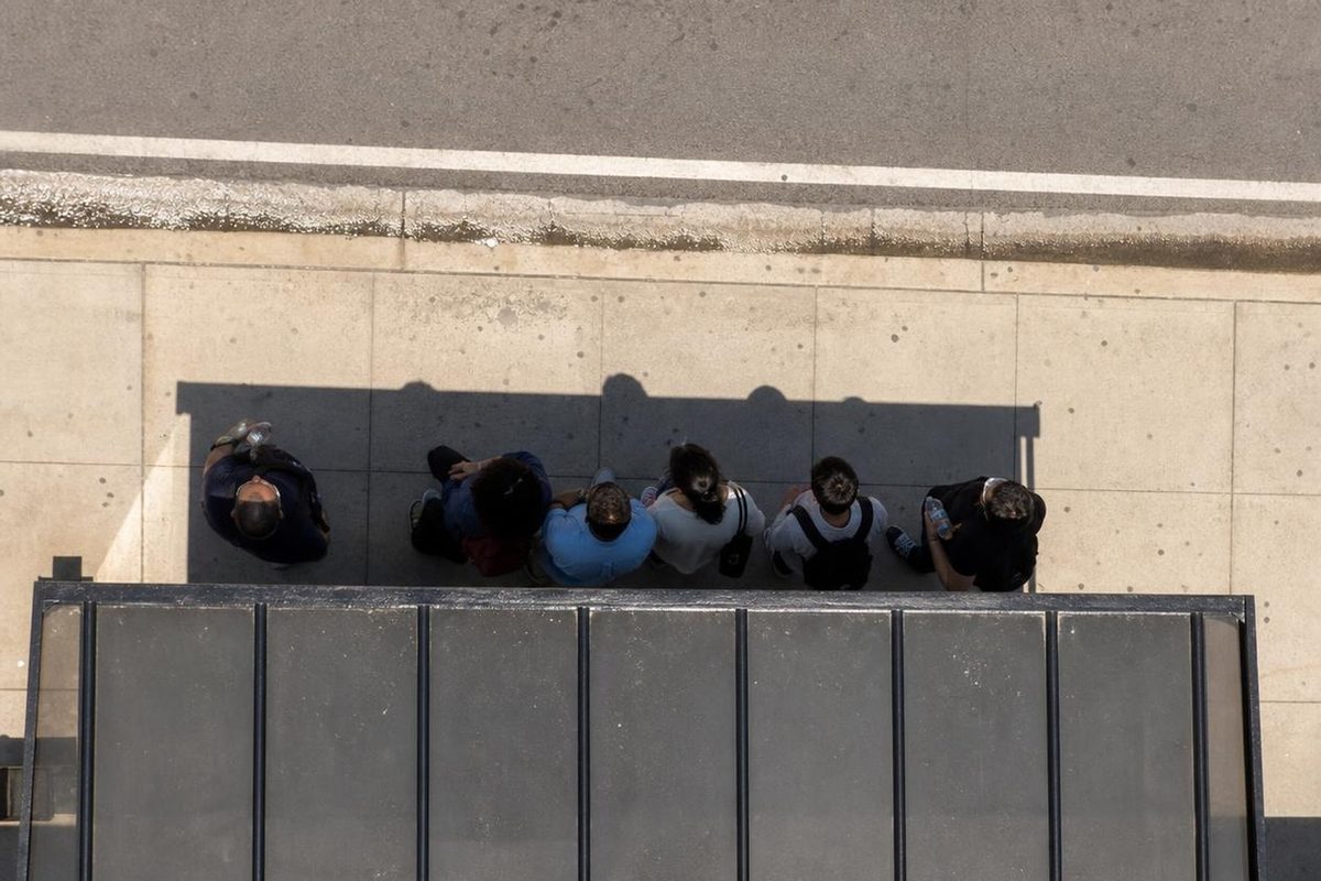 Overhead view of people crowded into the narrow shadow of a bus shelter on a sunbaked sidewalk at midday.