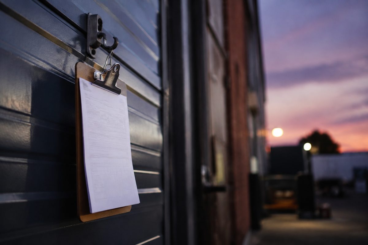 A blank call sheet clipped to a clipboard hanging beside a heavy soundstage door at dusk, the type unreadable and the background thrown out of focus.