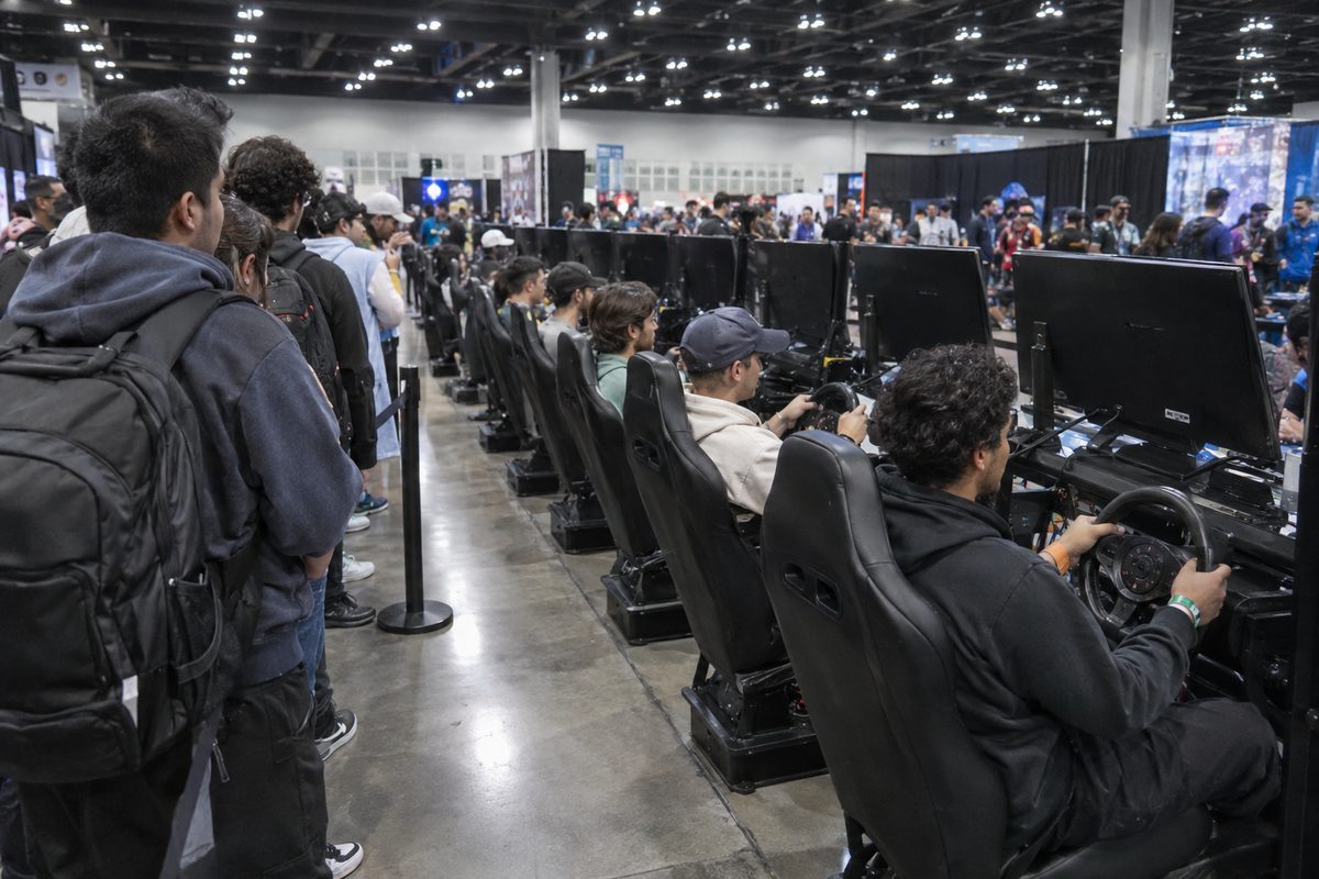 Wide view of a fluorescent-lit convention hall where people sit in racing seats with steering wheels at rows of game demo stations, seen from behind.