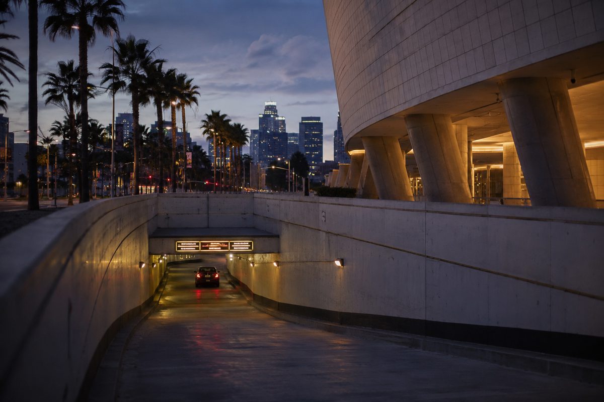 Photo illustration: the entrance ramp of a parking garage beneath a modern museum at dusk, a lit rate sign over the ramp and one car's tail lights descending, palm trees and the downtown Los Angeles skyline behind, no identifiable building, no text, no recognisable people
