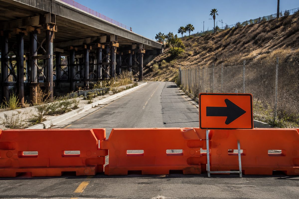 Orange barricades and a detour arrow block a two-lane surface street beside a freeway embankment under hard midday sun, with chain-link fencing and dry chaparral behind.