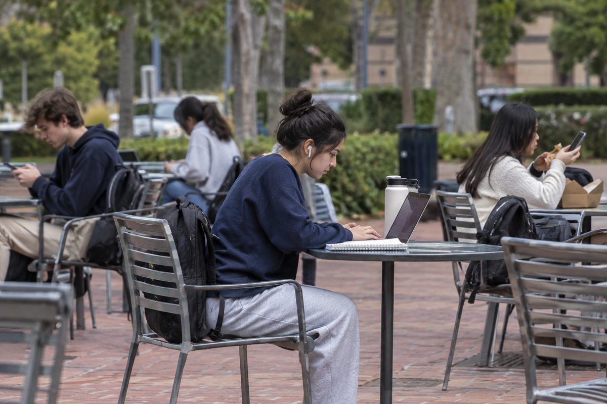 Students sitting at outdoor tables on a college campus on an overcast afternoon, typing on phones and laptops.