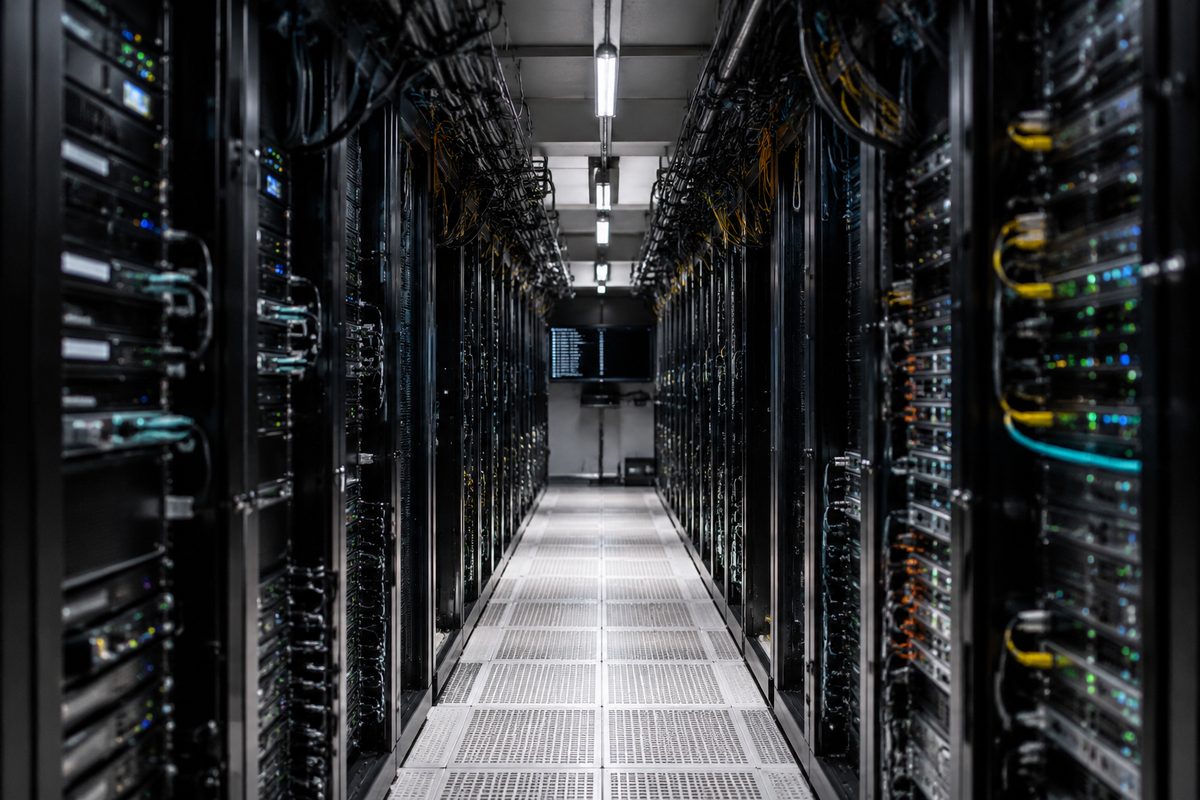 A frontal view of a fluorescent-lit server room aisle, cable trays overhead and rack fans in rows, with an unreadable terminal screen at the end of the aisle.