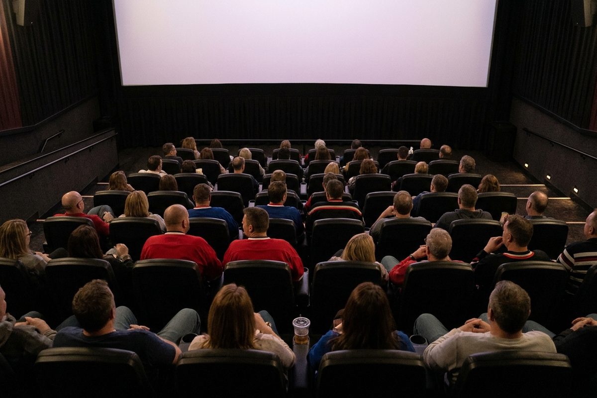Moviegoers, some in hockey jerseys, sit in a dark multiplex auditorium lit by the glow of the screen, seen from the back rows.