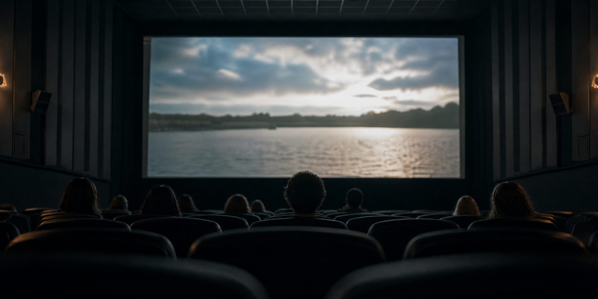 A symmetrical view down a darkened movie theater auditorium, rows of empty seats in the foreground and a few seated patrons silhouetted against the lit screen.