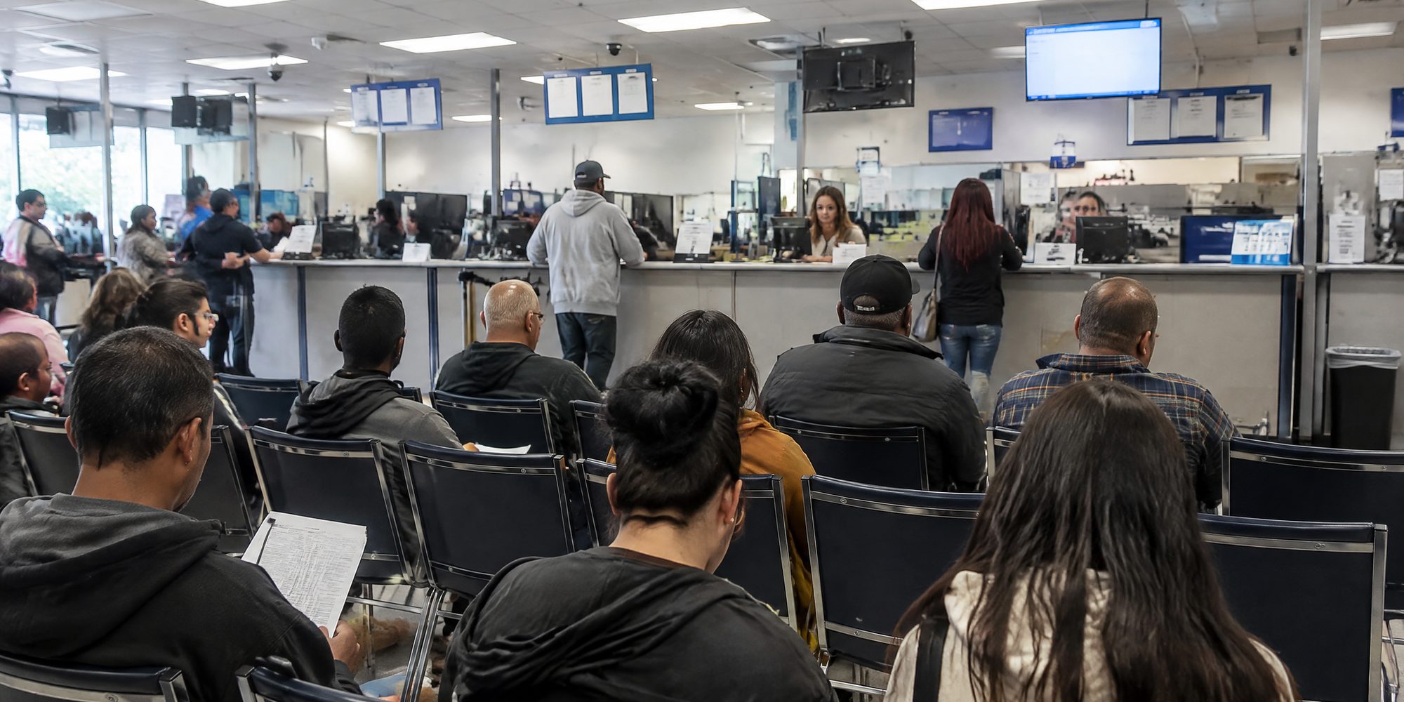 Rows of people sitting in a California DMV field office waiting area, seen from the back of the room, with service counters ahead under fluorescent light.
