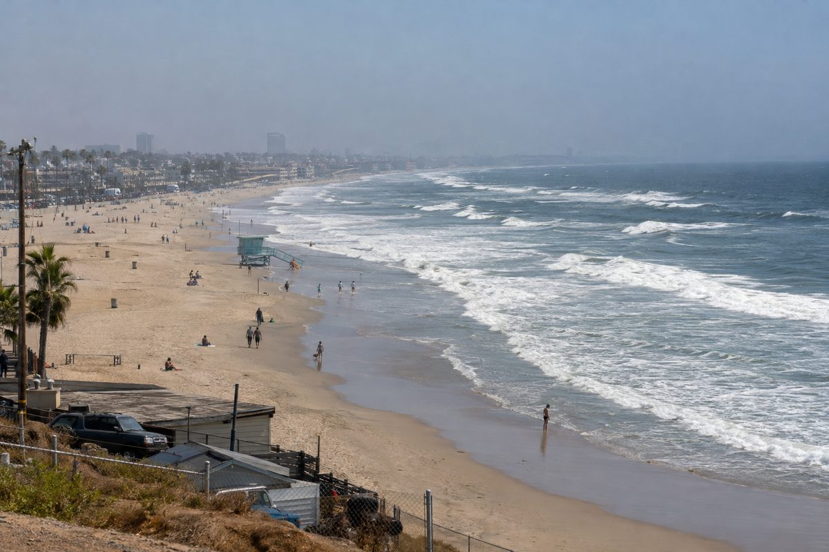 Waves breaking along a wide Southern California beach south of the Santa Monica Pier under a hazy afternoon sky, with a few people walking near a lifeguard tower as Hurricane Rachel swells arrive.