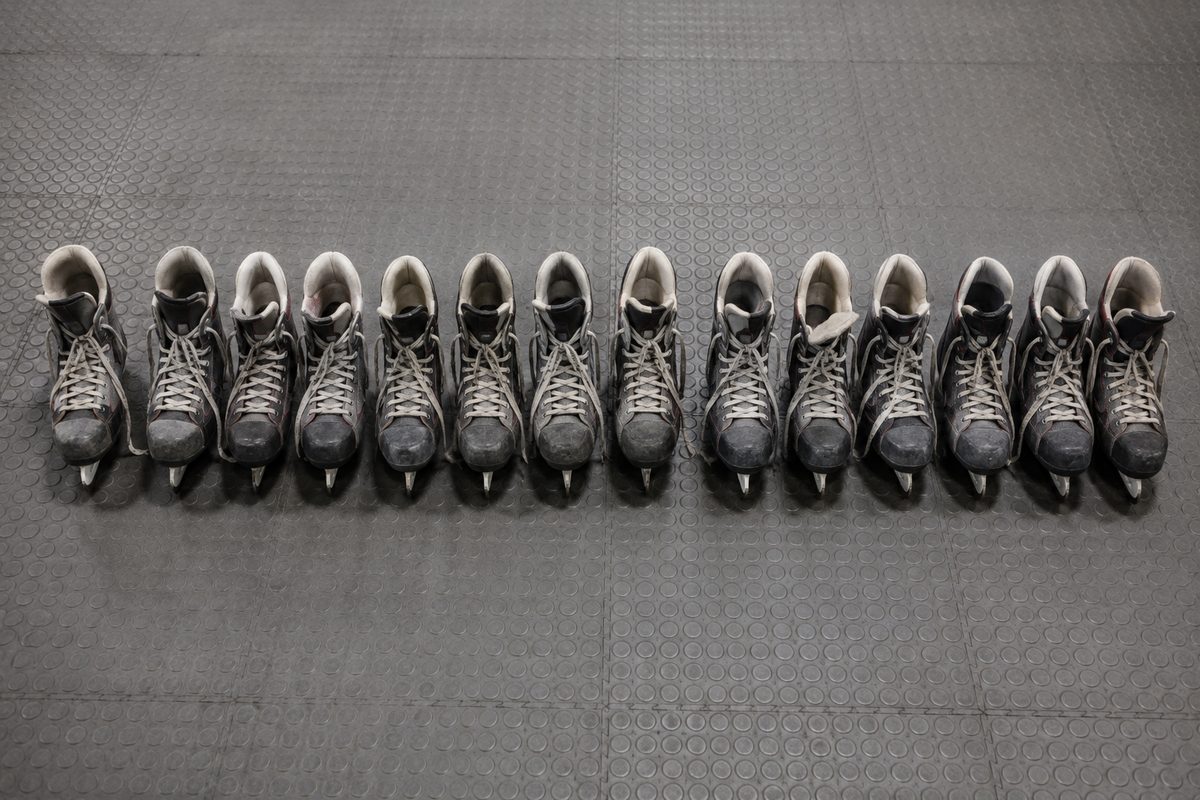 Photo illustration: a graphic overhead lineup of eleven pairs of well-worn rental ice skates placed in one straight row on a plain grey rubber floor, laces loose, no logos, no lettering, no people, hard even overhead light