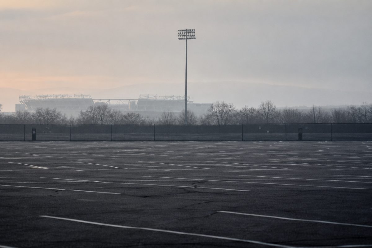 An empty asphalt parking lot at dawn, faded painted stall lines running to a chain-link fence, a light tower in silhouette against haze.