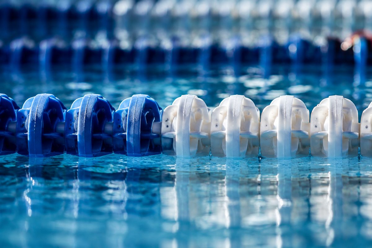 Photo illustration: extreme macro of blue-and-white lane ropes floating on still indoor pool water, hard overhead light, no swimmers, no signage, no venue identifiers