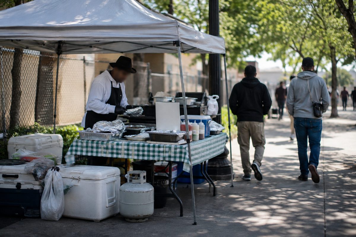 A folding-table food vending setup under a canopy on a sunlit city sidewalk, with hands wrapping food and pedestrians walking past.