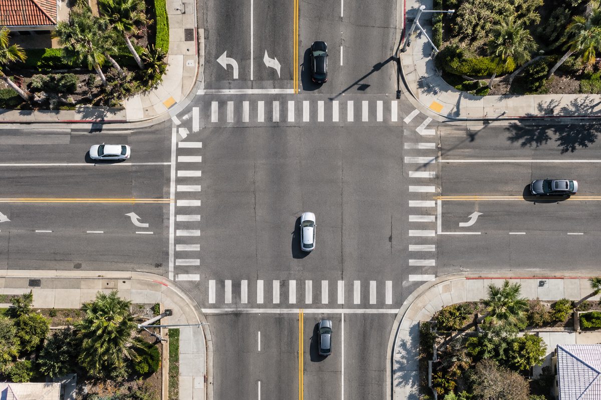 Overhead view of a wide suburban boulevard intersection at midday, crosswalk stripes and turn arrows painted on empty asphalt.