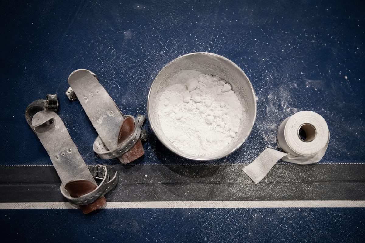 Overhead view of a chalk bowl, gymnastics hand grips and wrist tape arranged on a gym floor under night lighting.