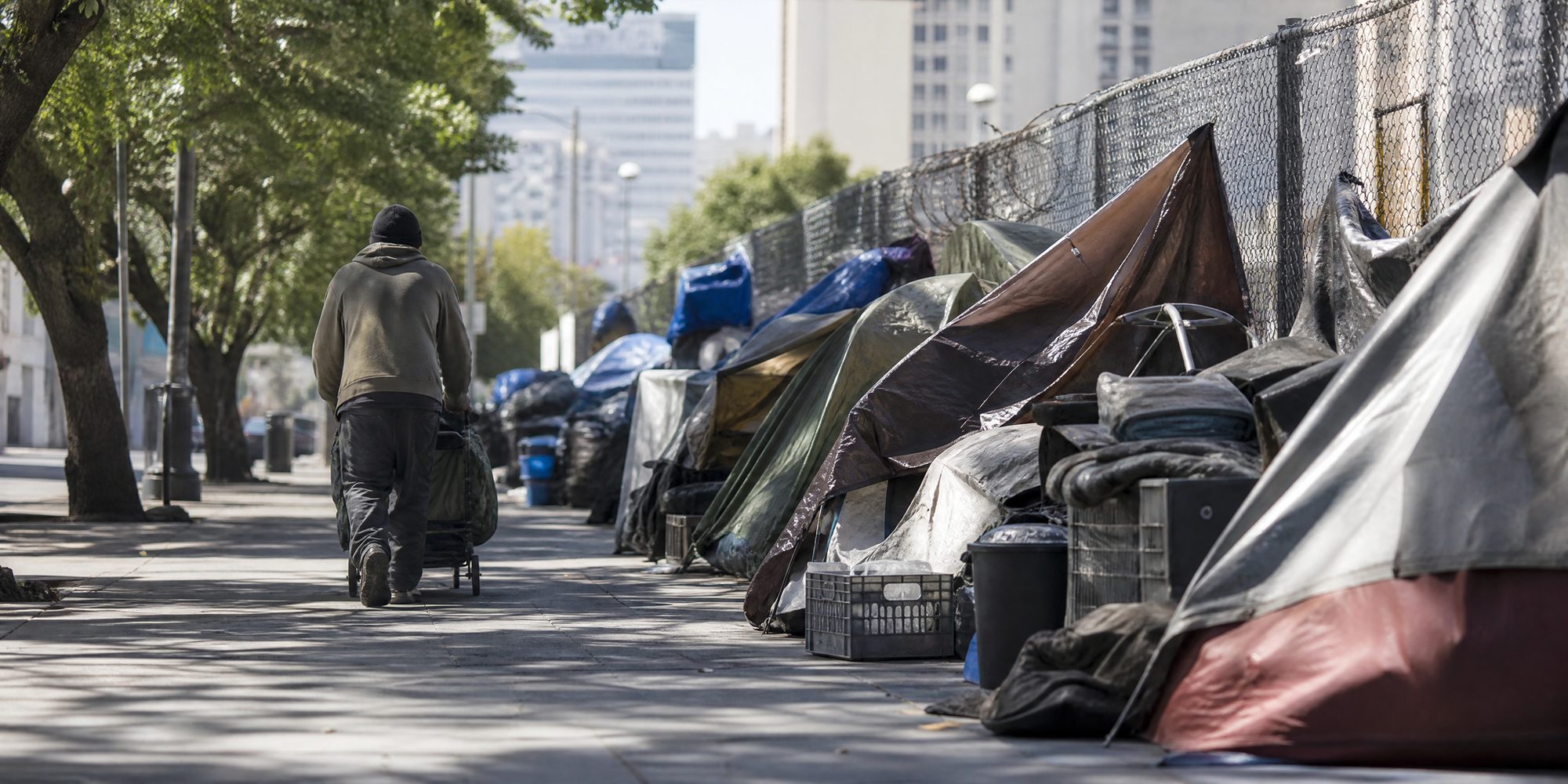 A downtown sidewalk at midday with tents, tarps and stacked belongings against a chain-link fence, a person walking away from the camera pushing a cart.