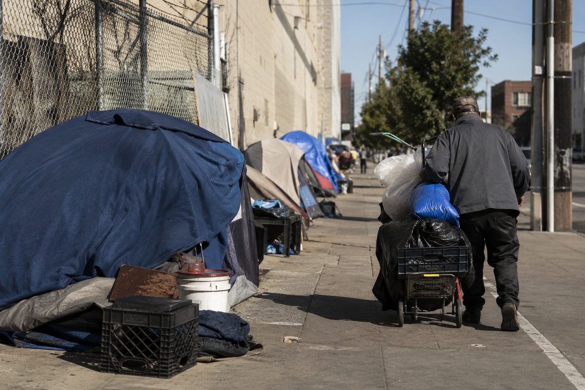 A downtown sidewalk at midday with tents, tarps and stacked belongings against a chain-link fence, a person walking away from the camera pushing a cart.