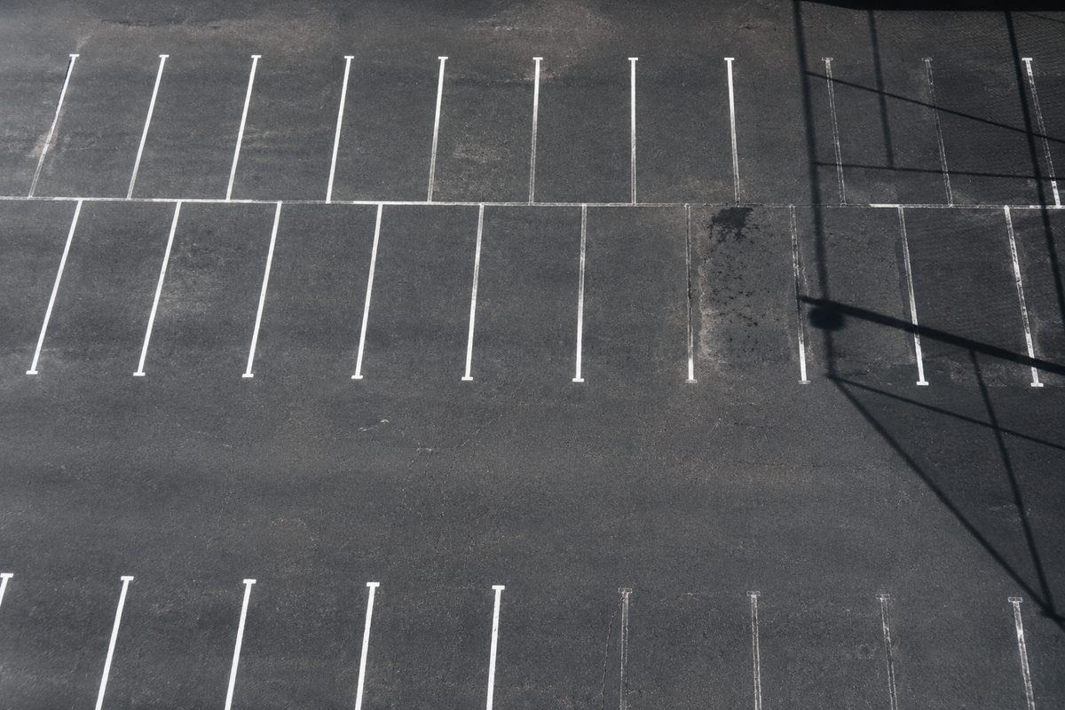 Overhead view of an empty asphalt lot in hard noon light, its parking stalls marked in faded white paint, with several spaces unmarked.