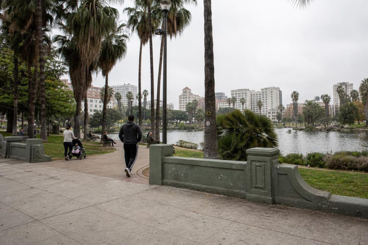 People walk the paths beside the lake at MacArthur Park in Los Angeles on an overcast morning, with Westlake buildings behind the trees.