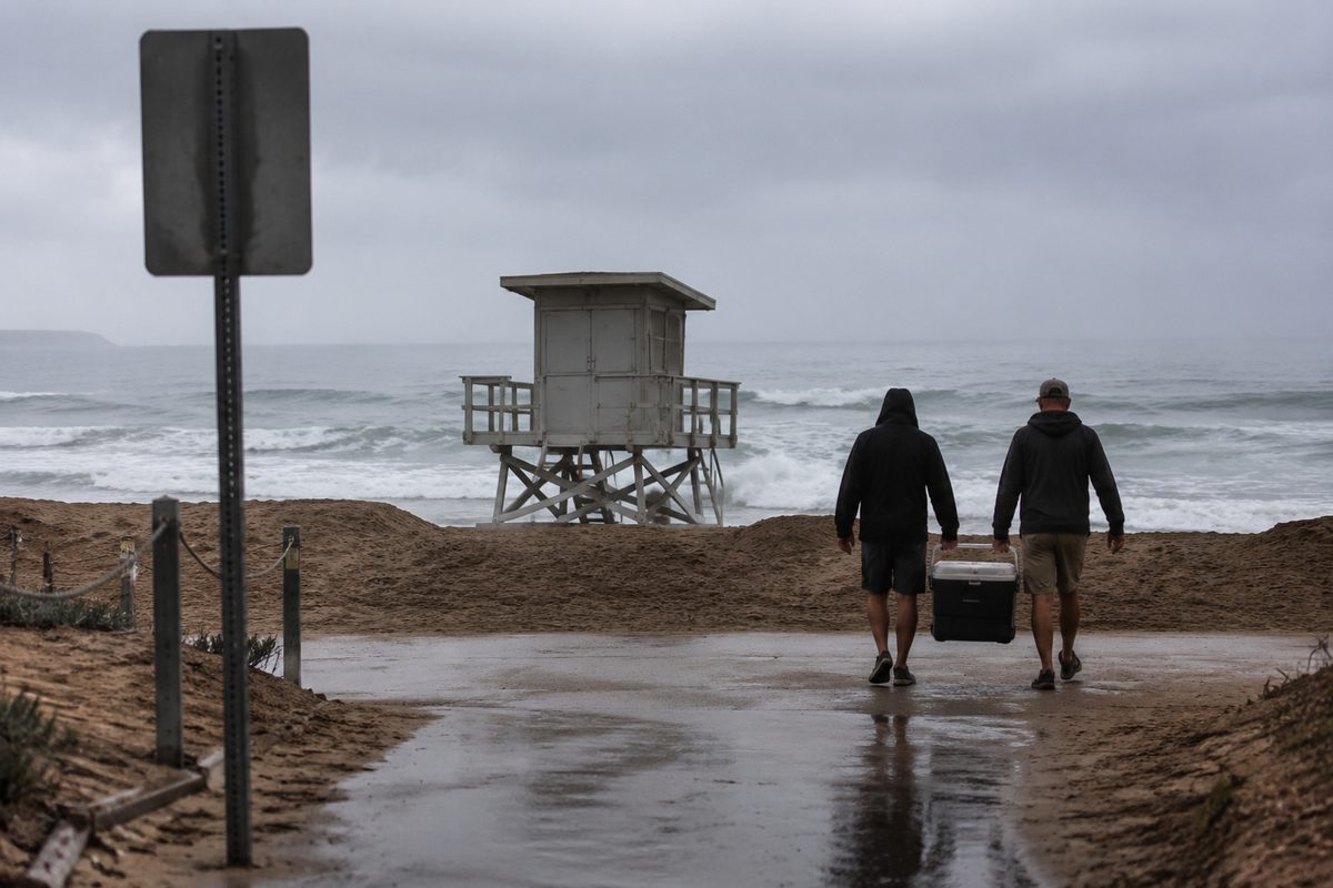 Marine-layer morning on a beach path, figures seen from behind at the top of a sand berm, a shuttered lifeguard tower mid-frame and foam pushing across dark wet sand.