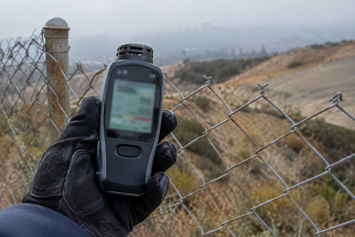 A gloved hand holds a handheld air monitor beside a chain-link fence, with a dry hillside fading into morning haze behind it.