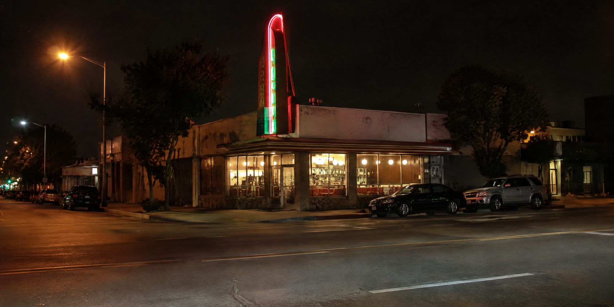 Photo illustration: A classic old-school Los Angeles deli at night in the Fairfax District, its tall vertical neon sign glowing red and green above the storefront over a wide, nearly empty city avenue, seen from across the street at sidewalk level, lit only by the neon, the deli's own window lights and orange streetlamps, a few parked cars at the curb, late-night quiet, no people, and no readable lettering, logos or prices anywhere in the frame.