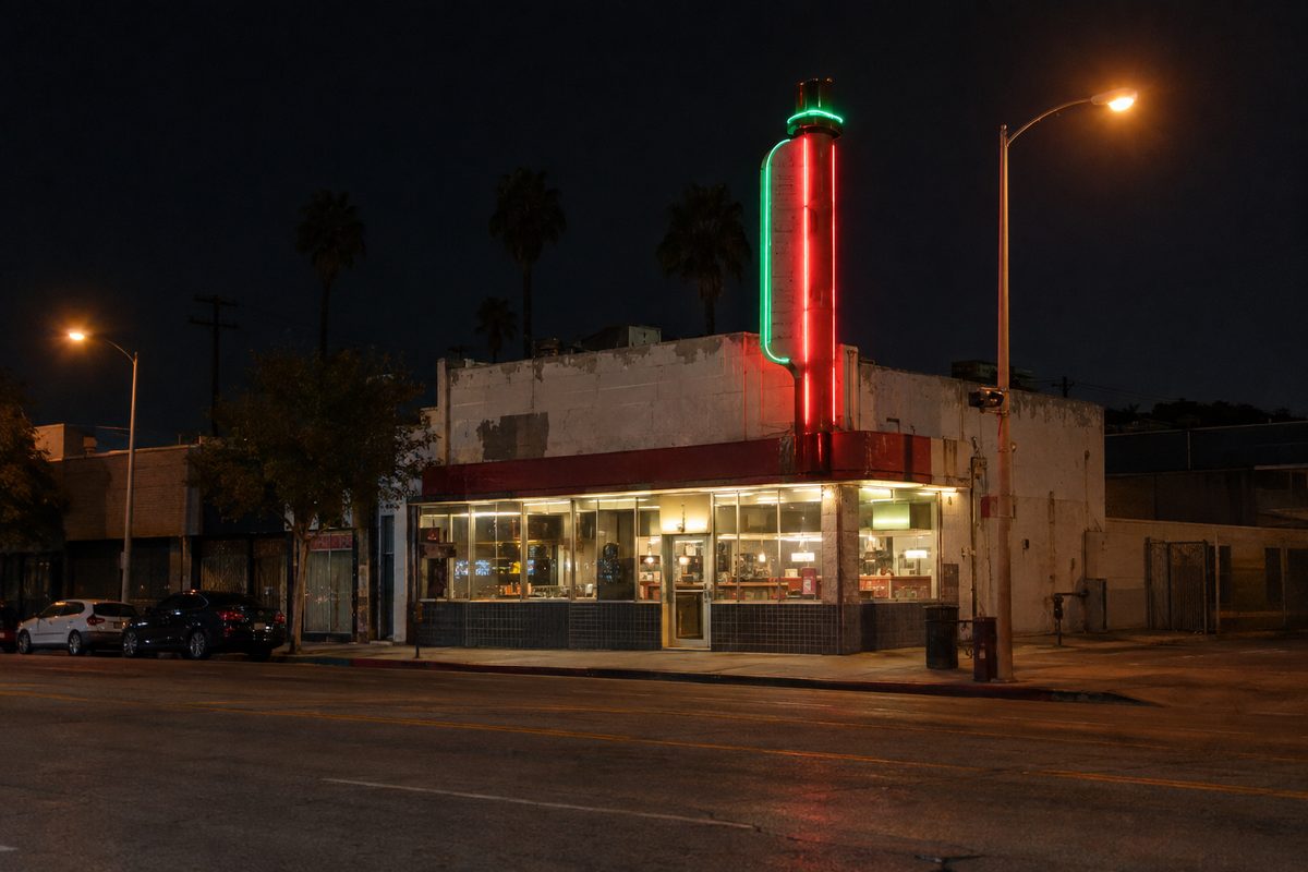 Photo illustration: A classic old-school Los Angeles deli at night in the Fairfax District, its tall vertical neon sign glowing red and green above the storefront over a wide, nearly empty city avenue, seen from across the street at sidewalk level, lit only by the neon, the deli's own window lights and orange streetlamps, a few parked cars at the curb, late-night quiet, no people, and no readable lettering, logos or prices anywhere in the frame.