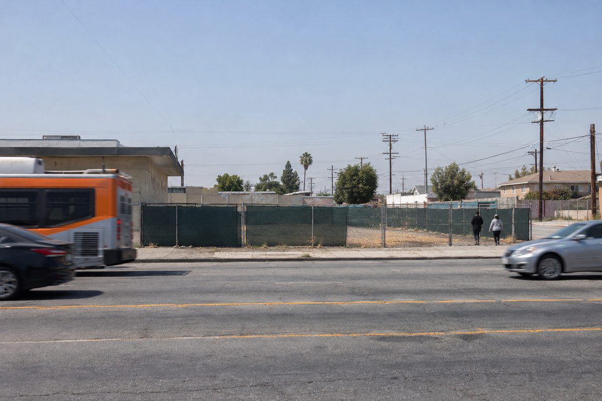 A chain-link construction fence with green screening runs along the sidewalk of a commercial stretch of South Vermont Avenue in Vermont Square, South Los Angeles, with storefronts beyond.