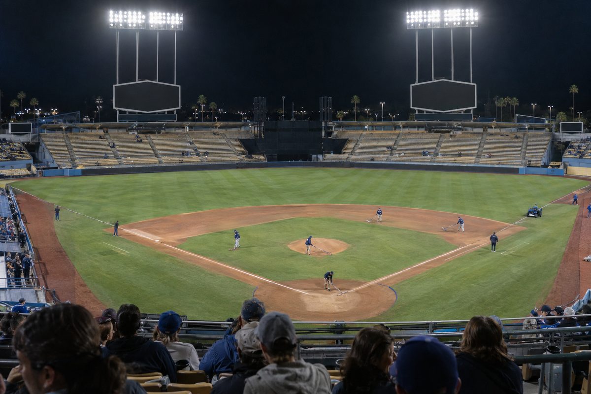 Photo illustration: Last night in Los Angeles sports: the field of a Los Angeles baseball stadium just after a night game, the floodlights still on over the emptying outfield seats and a grounds crew raking the infield dirt, seen from the upper deck behind home plate.