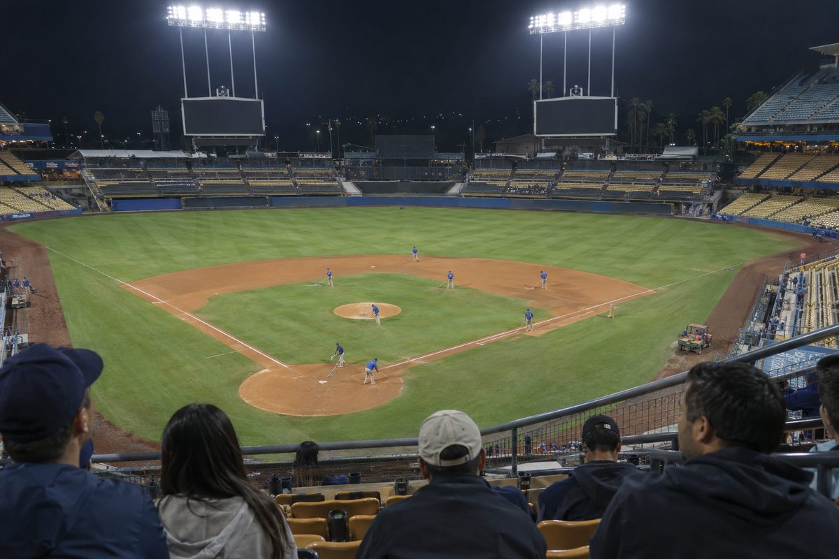 Photo illustration: Last night in Los Angeles sports: the field of a Los Angeles baseball stadium just after a night game, the floodlights still on over the emptying outfield seats and a grounds crew raking the infield dirt, seen from the upper deck behind home plate.