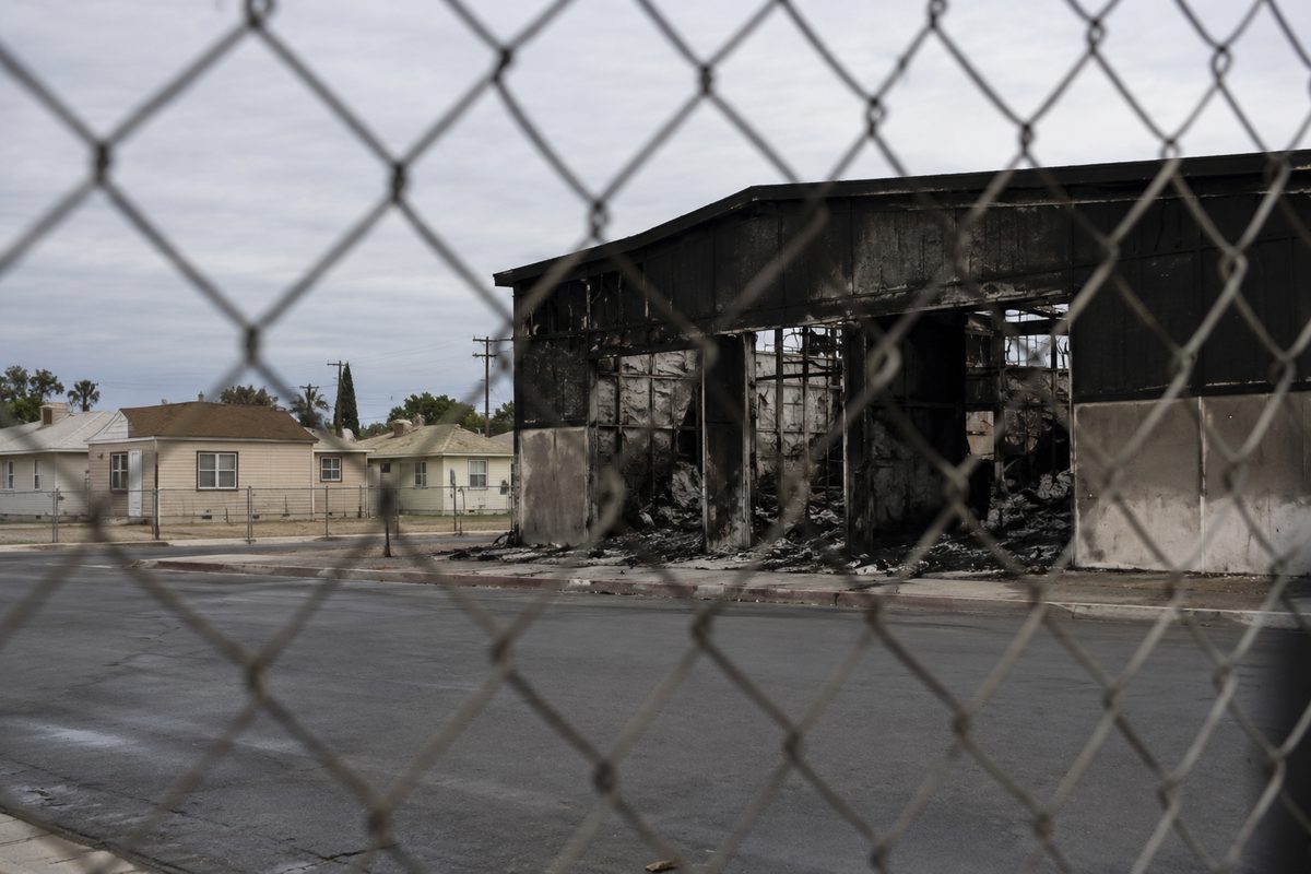 Chain-link fence in front of a burned, boarded industrial warehouse, with single-story houses visible across the street.
