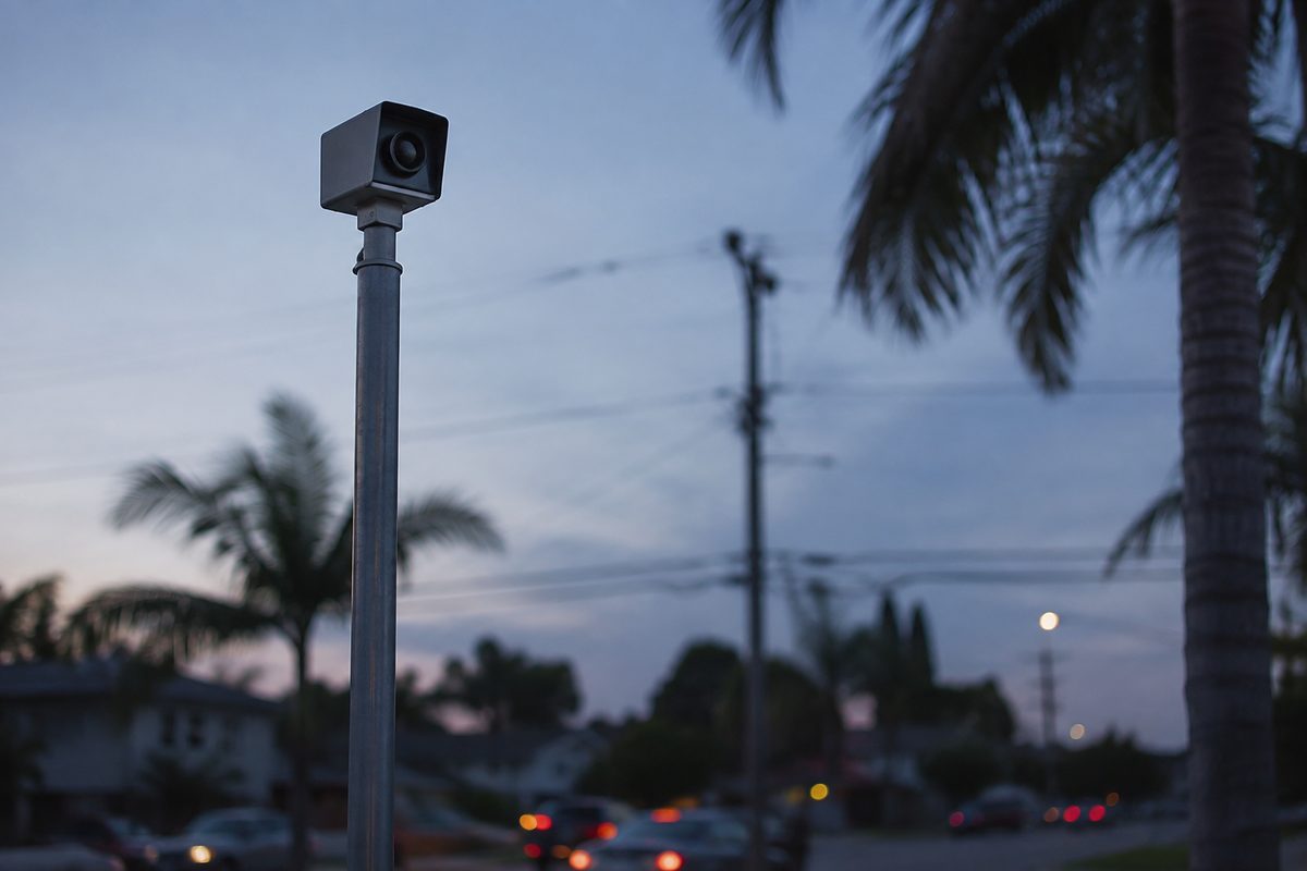 A small white surveillance camera on a metal pole above an unnamed residential street, angled toward passing traffic at dusk.