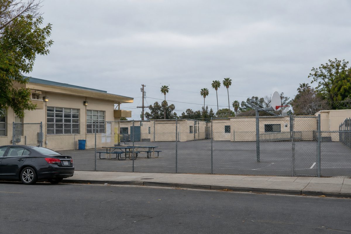 Photo illustration: The street front of a public elementary school campus in Los Angeles on an overcast weekday morning, seen from the sidewalk across the street: low beige stucco classroom buildings with a row of portable classrooms behind a tall chain-link fence, a mostly empty asphalt yard with a few lunch tables and a basketball hoop, a parked car or two at the curb, flat gray sky and even light, no people in the frame, and no text, signs, prices or logos anywhere in the picture.