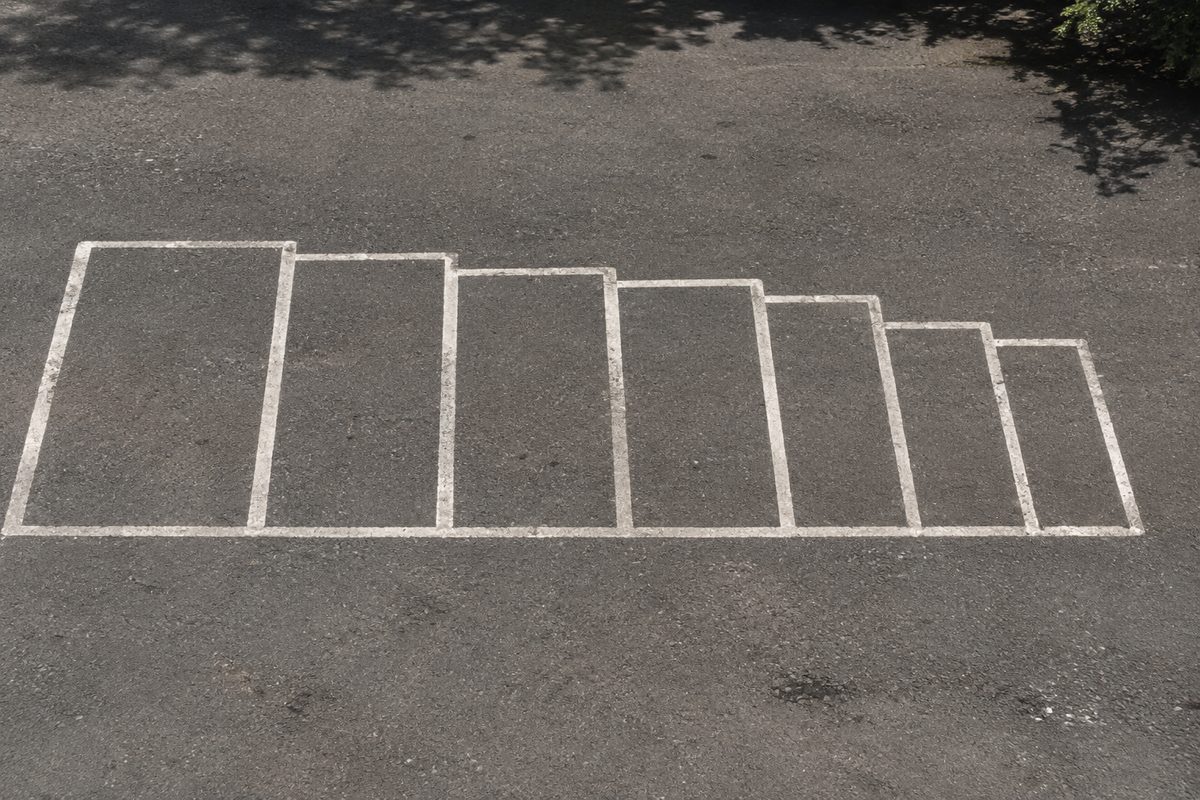 Photo illustration: a graphic overhead lineup of eight blank parking-space rectangles painted on asphalt in one straight row, each stall progressively smaller than the last, hard noon light, no cars, no signage, no logos, no readable text