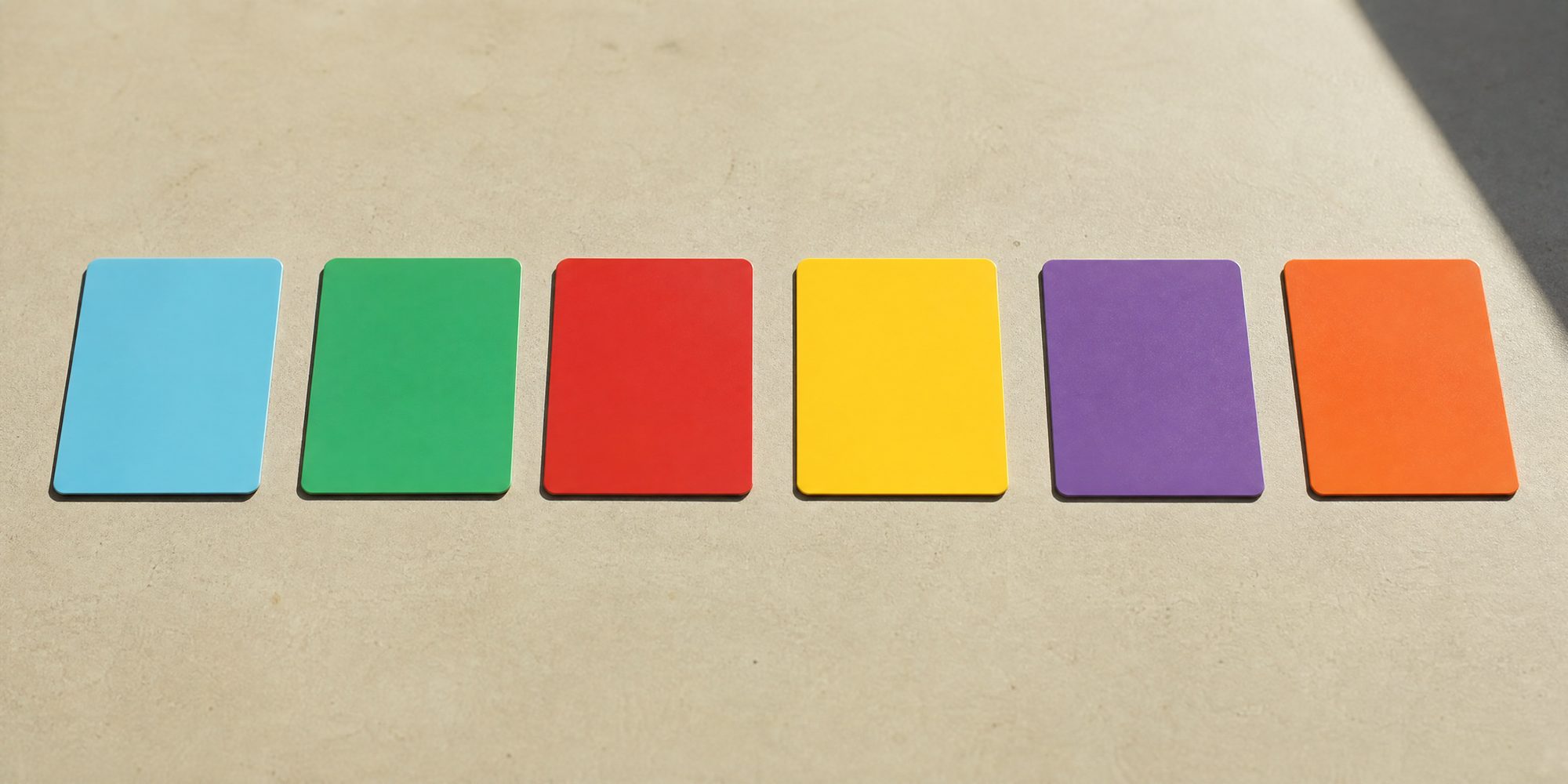 Photo illustration: a graphic overhead lineup of six blank rectangular plastic cards laid in one straight row on a plain table, each a different flat colour, no text, no logos, no lettering, no hands, hard noon light