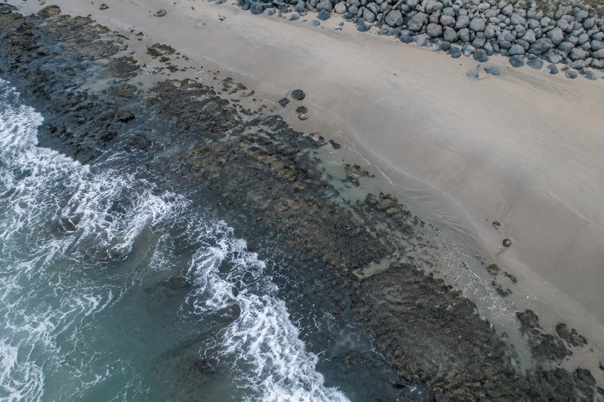 Overhead view of a shoreline in morning haze: a dark rocky intertidal shelf meeting pale dry sand, with a line of stacked boulders along the upper edge.