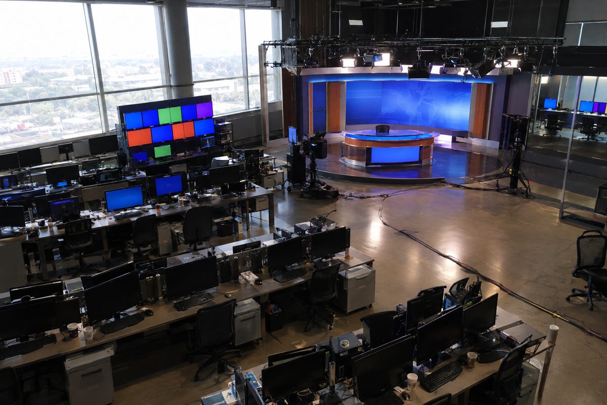Photo illustration: The newsroom of a national television news network on an ordinary weekday morning, seen wide from the back of the room: rows of desks with computer monitors and keyboards, a wall of broadcast screens glowing with plain color fields, an empty anchor desk under the room's own overhead studio lights, cables taped along the floor, a few coffee cups left at workstations, no people, and no text, prices or logos anywhere in the frame.