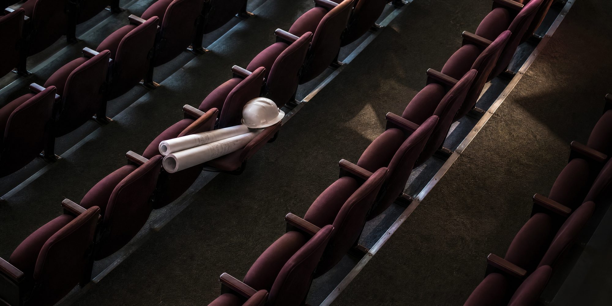 Overhead view of rows of theater seats in an unnamed auditorium, with rolled plans and a hard hat left on one seat in cool morning light.