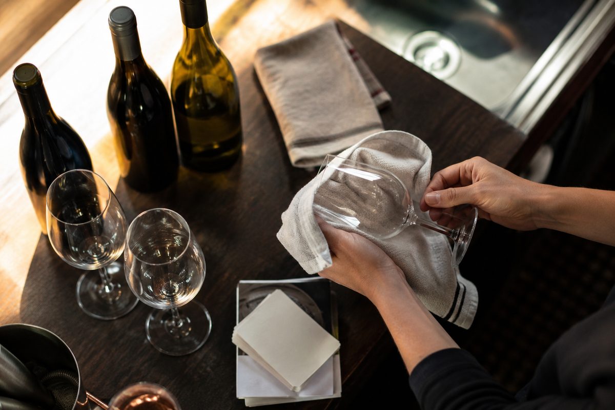 Overhead view of a bar service station with wine bottles, empty stemware, a folded towel and hands drying a glass, illustrating drink prices at a new Arts District rooftop with movie screenings.