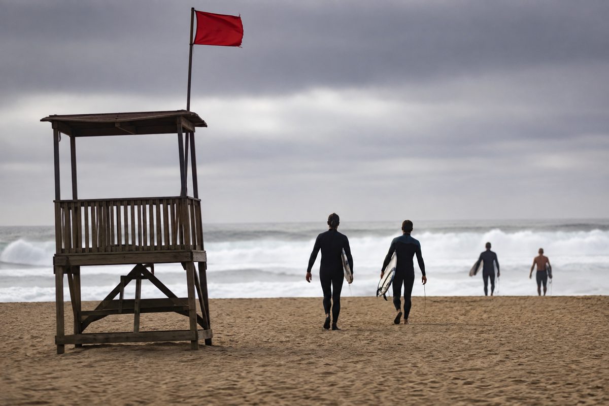 An unmarked lifeguard tower above an empty stretch of sand, gray marine layer overhead, surf breaking in the background as a few people walk toward the water.