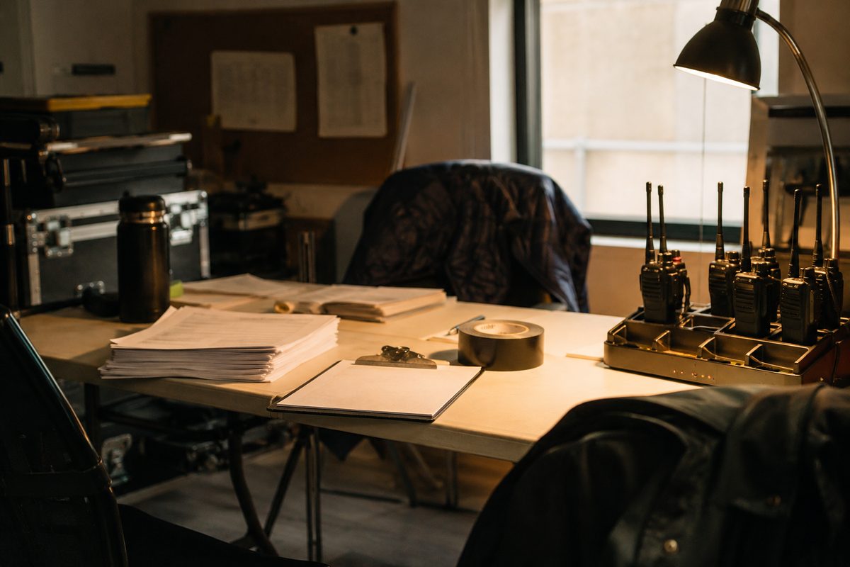 A folding table in a dim production office holding a stack of unmarked permit paperwork, a clipboard, a roll of gaffer tape and a charging bank of radios.