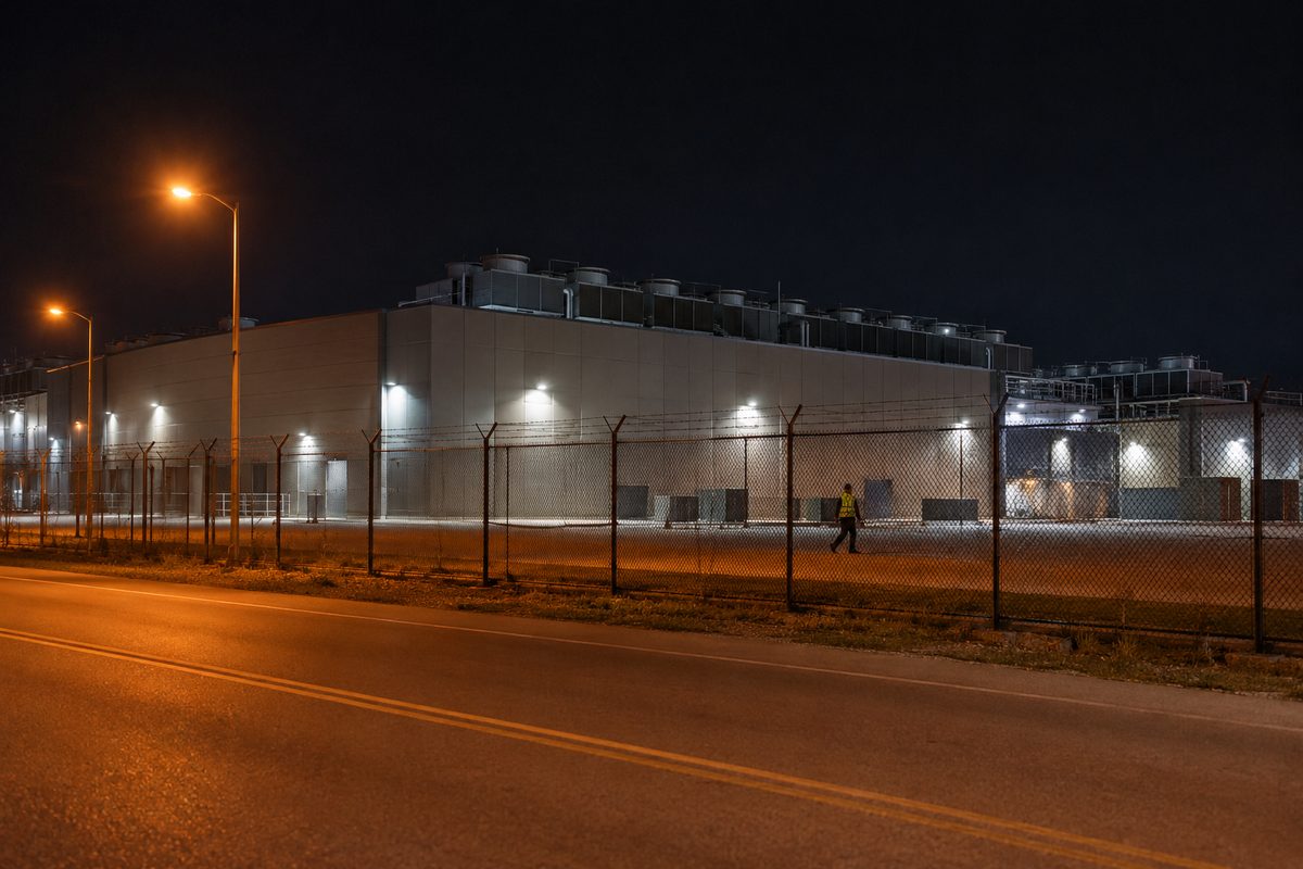 A large windowless data center building behind a chain-link fence at night, lit by streetlights along an empty access road.