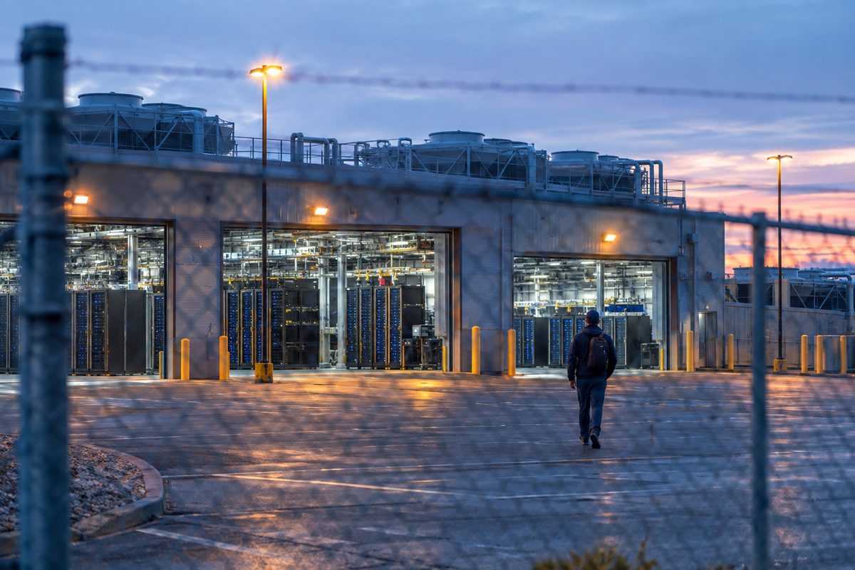 Rows of server racks seen through a chain-link fence at dawn, compressed by a long lens, with a technician walking away across an empty parking lot.