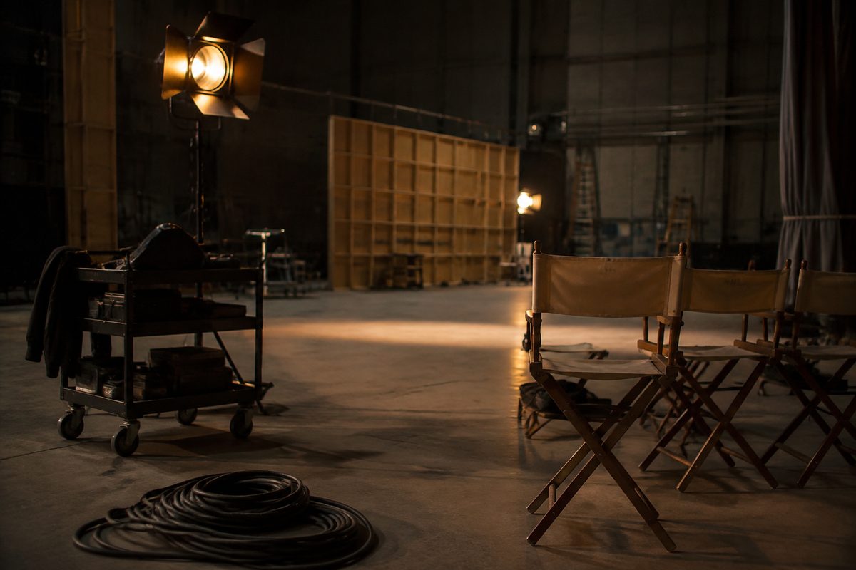 Wide view of an empty soundstage interior lit by warm work lamps, with coiled cable, a wheeled equipment cart and folded canvas chairs.