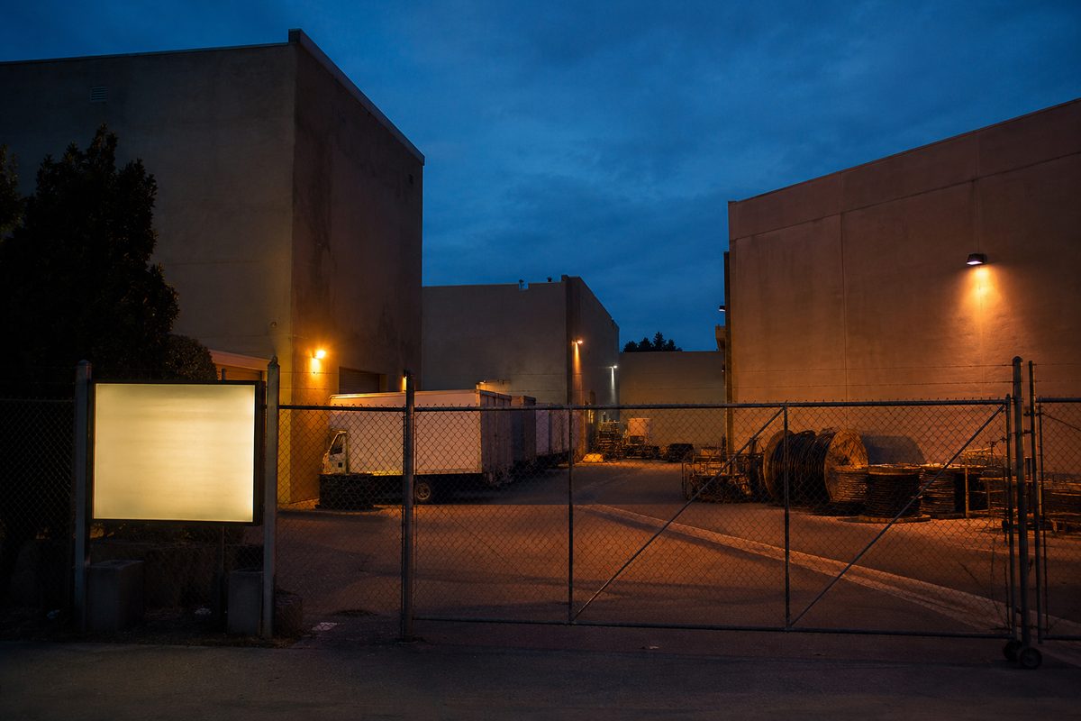 Wide dusk view of an anonymous studio backlot: blank soundstage walls, a chain-link gate and parked equipment trucks under sodium lights.