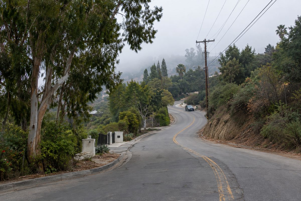 A winding hillside road in Benedict Canyon, Los Angeles, under an overcast morning sky, tied to the Manson family's Tate-LaBianca murders.