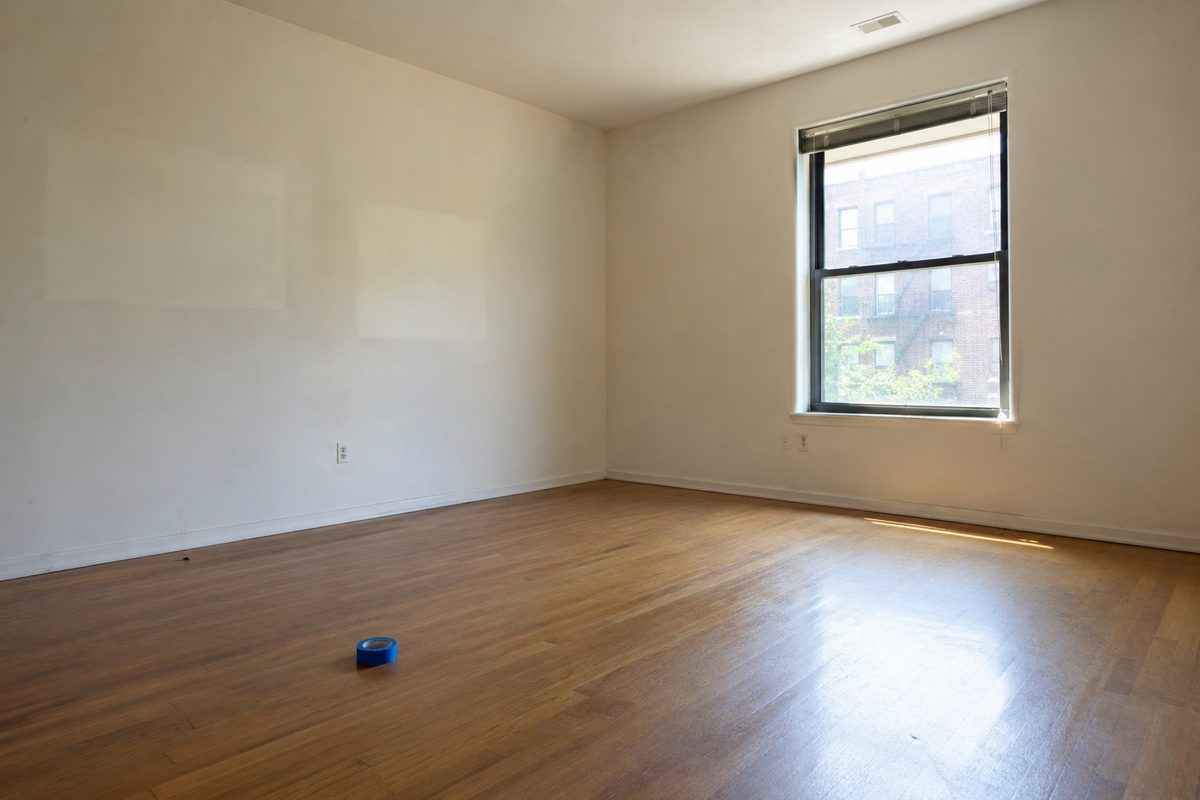 Photo illustration: an empty rental apartment on move-out day, bare wood floor, blank white walls with faint rectangles where pictures hung, one window with no curtain, afternoon light, a single roll of blue painter's tape on the floor, no people, no furniture, no signage, no addresses