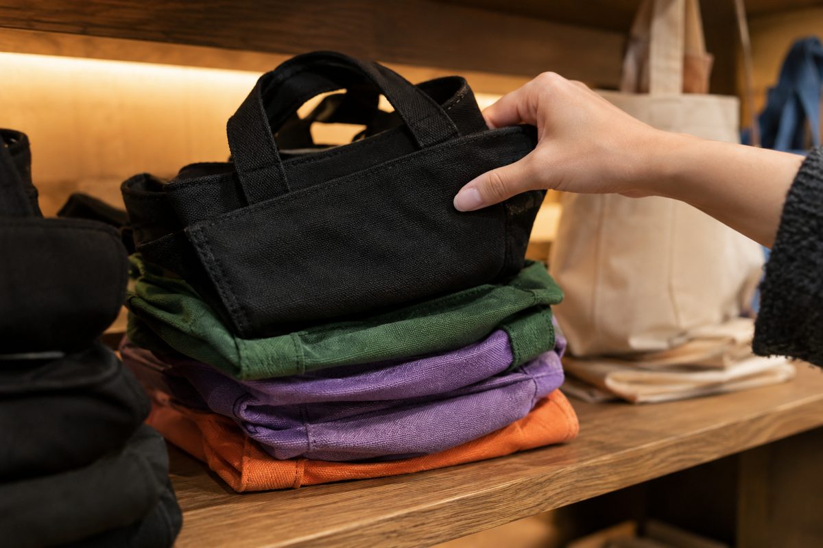 Close-up of small canvas tote bags in black, green, purple and orange stacked on a wooden shelf as a hand lifts one.